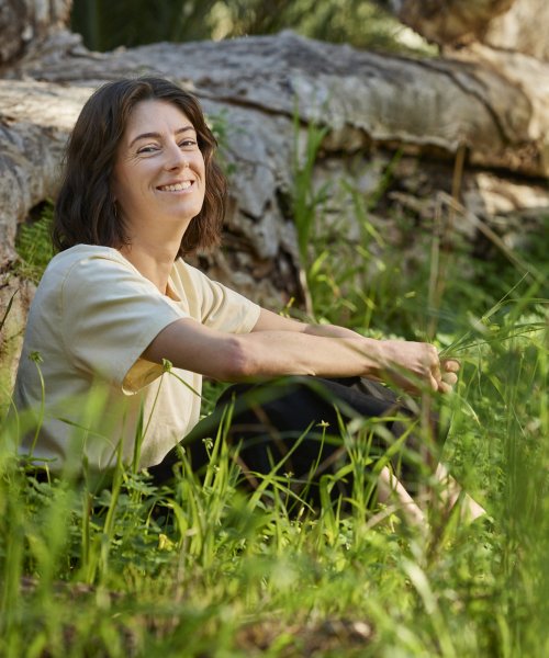 A happy woman smiles while seated in the grass near a large fallen tree trunk
